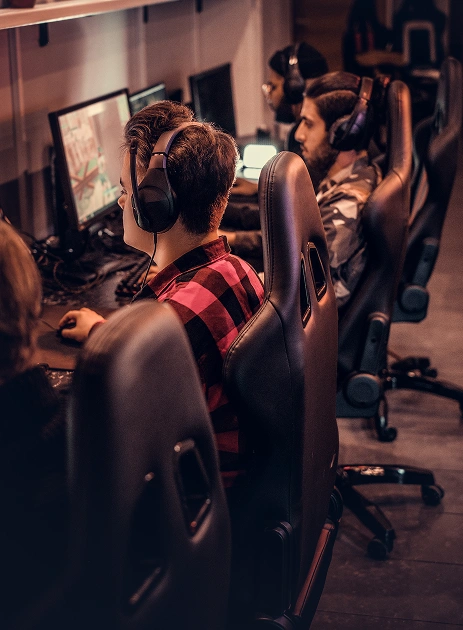 Uma fileira de jovens sentados em cadeiras de jogo pretas e usando fones de ouvido enquanto jogam focados em uma lan house ou arena gamer.